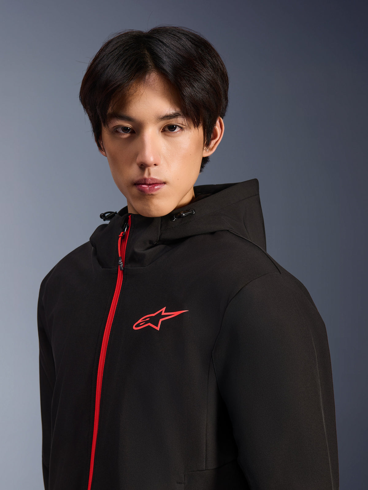 Alpinestars Radium Tech Hoodie, veste de moto urbaine, noir et rouge vif, modèle masculin portant un sweat à capuche en coque technique avec une fermeture éclair frontale et un logo sur la poitrine rouges, design de conduite urbaine moderne