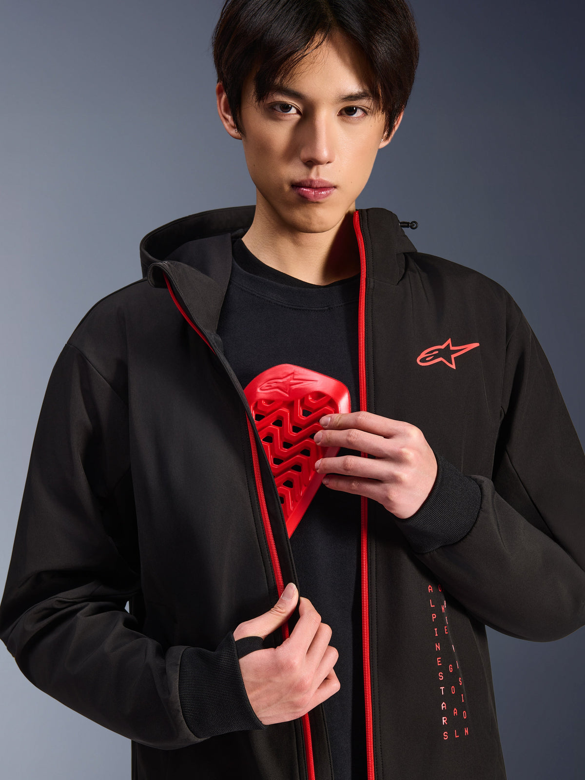 Alpinestars Radium Tech Hoodie, veste de moto urbaine, noir et rouge vif, porté par un mannequin montrant un insert de protection pectorale rouge, doté de fermetures éclair rouge vif et d'un marquage Alpinestars vertical, coque technique coupe-vent épurée pour les trajets urbains