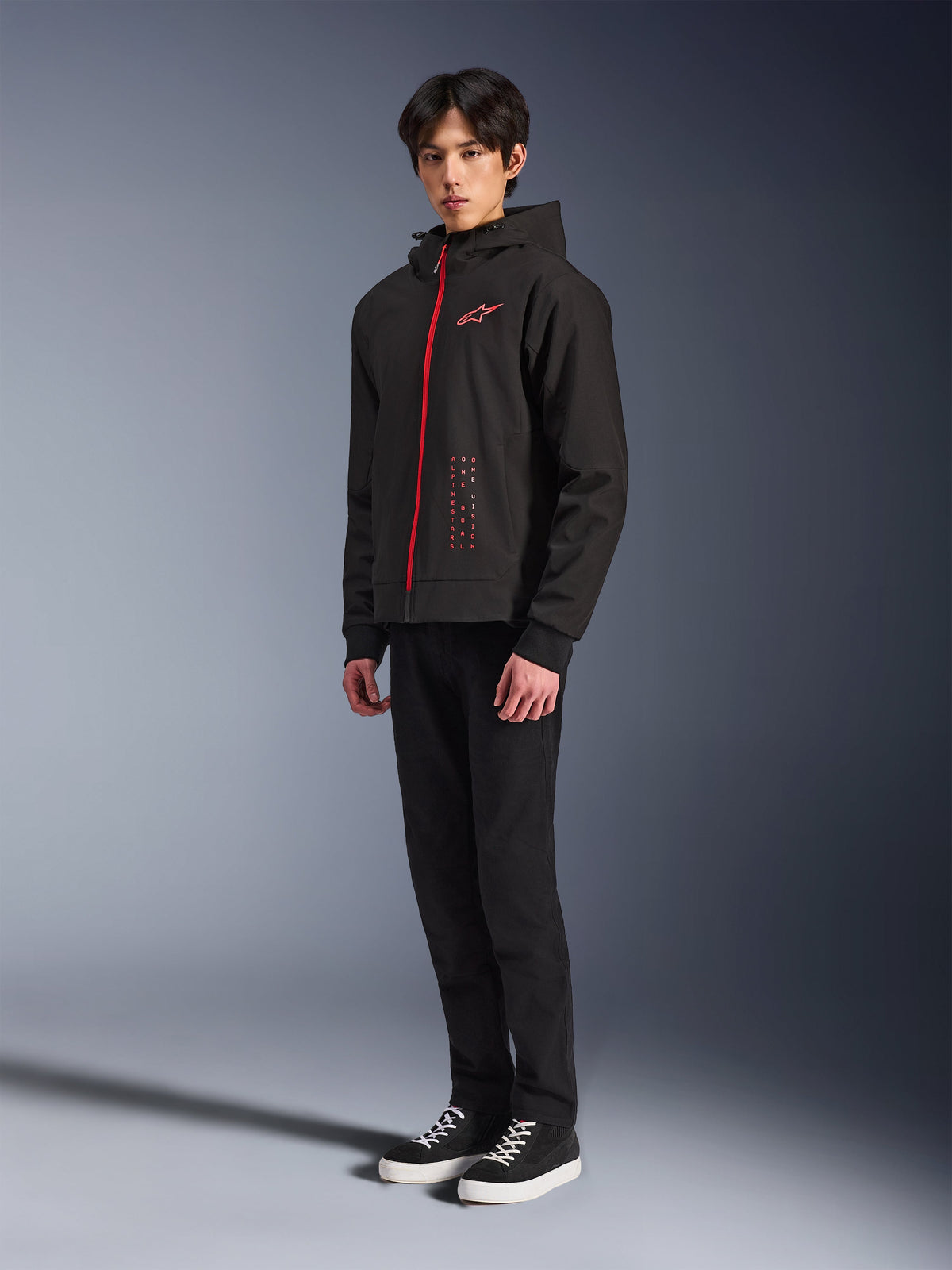 Alpinestars Radium Tech Hoodie, Noir Rouge Vif, sweat à capuche technique pour la conduite urbaine doté d'une fermeture éclair frontale rouge et d'un logo sur la poitrine, porté par un mannequin avec un pantalon noir et des baskets noires à semelles blanches, photo de studio en pied