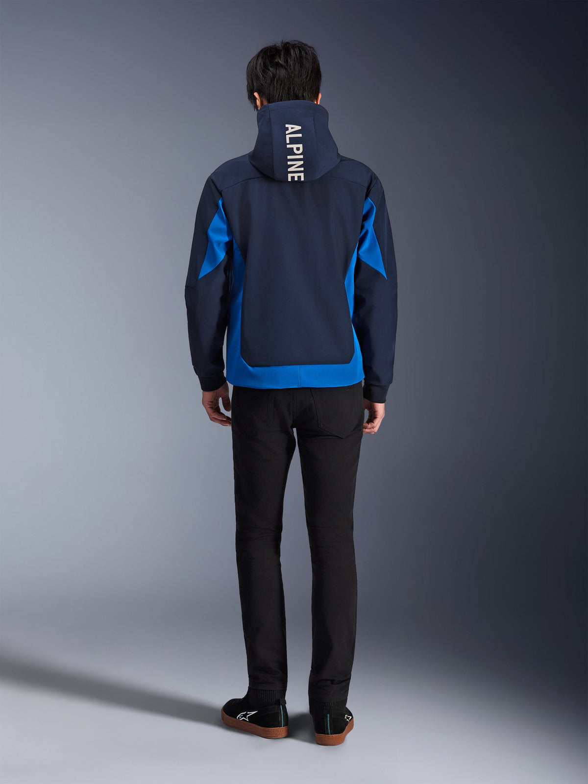 Hoodie Tech Radium Alpinestars, bleu marine et bleu royal, vue de dos montrant le marquage vertical « ALPINE » sur la capuche, panneaux latéraux et ourlet bleu royal, hoodie de moto urbain décontracté avec coque 3L coupe-vent, porté avec un pantalon noir