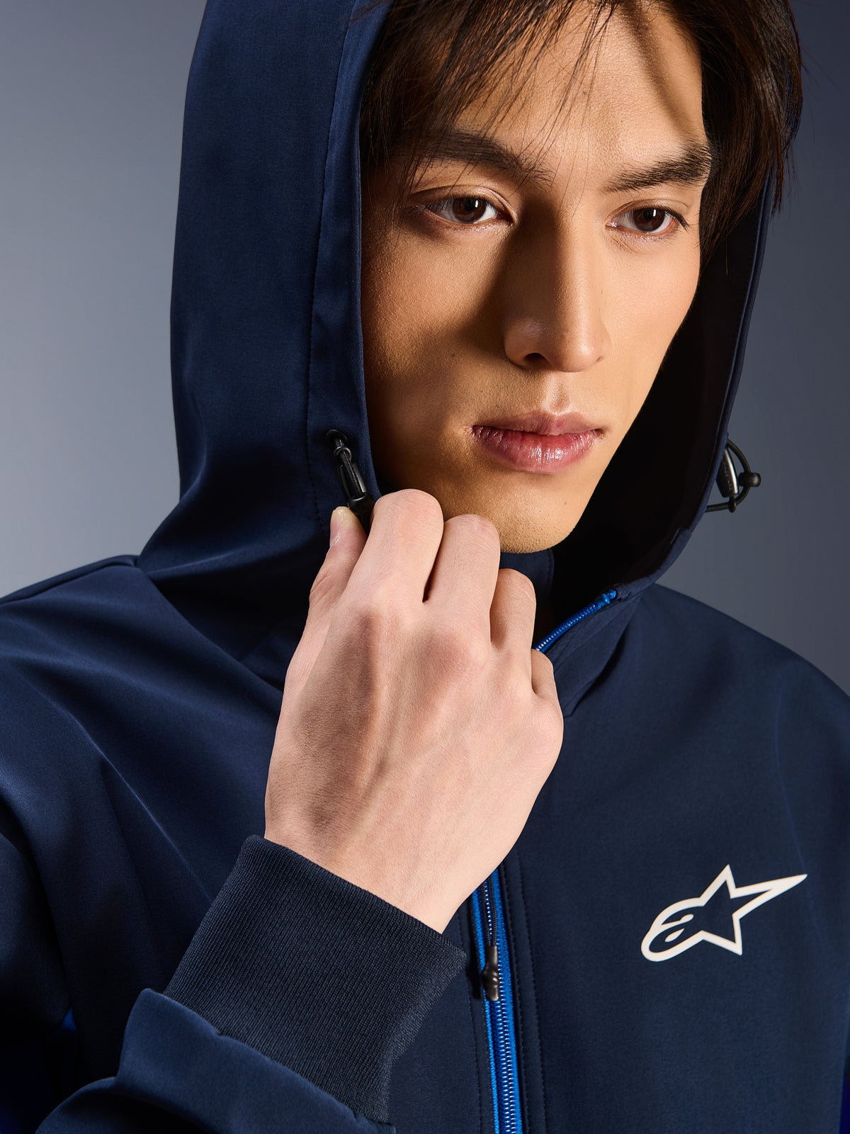 Alpinestars Radium Tech Hoodie, Night Navy Royal Blue, gros plan d'un homme portant le hoodie avec la capuche relevée, logo Astars blanc sur la poitrine, fermeture éclair bleue, veste technique urbaine