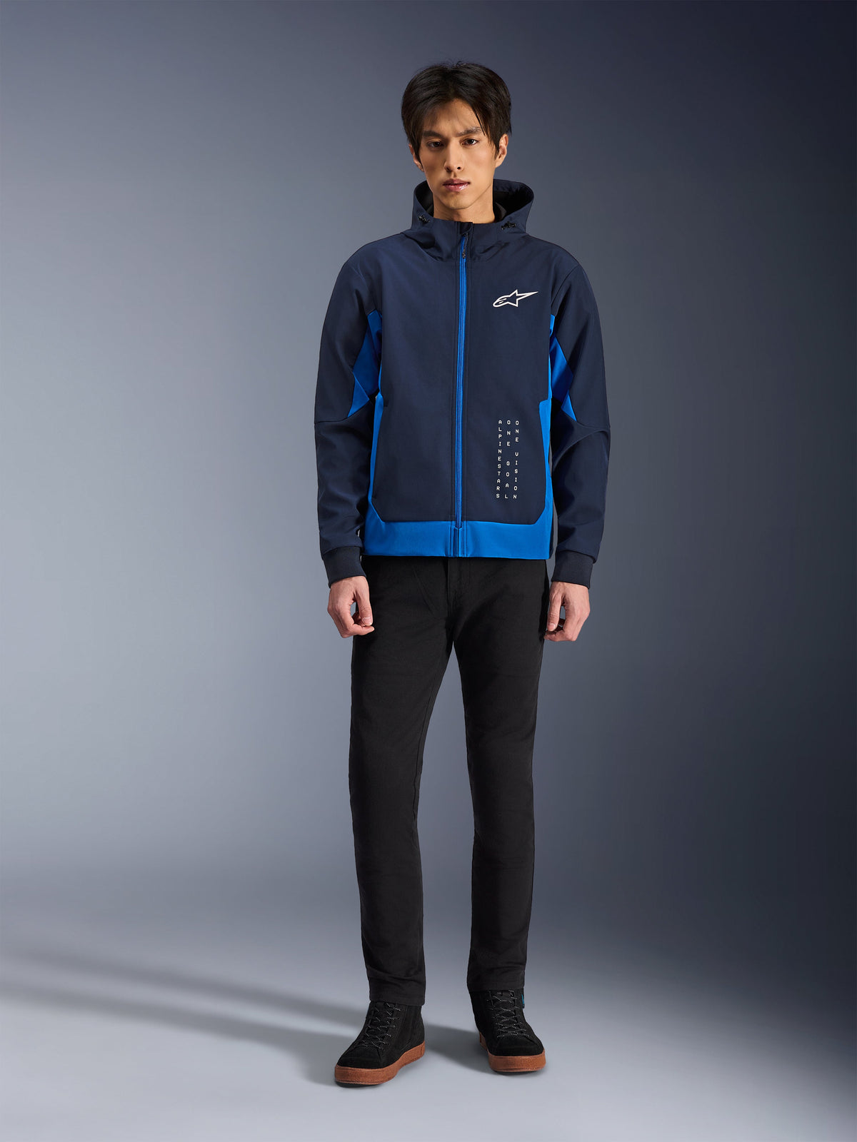 Alpinestars Radium Tech Hoodie, sweat à capuche de moto urbain, bleu marine et bleu royal, coque technique coupe-vent, design streetwear décontracté avec logo blanc sur la poitrine, porté avec un pantalon noir et des baskets montantes.