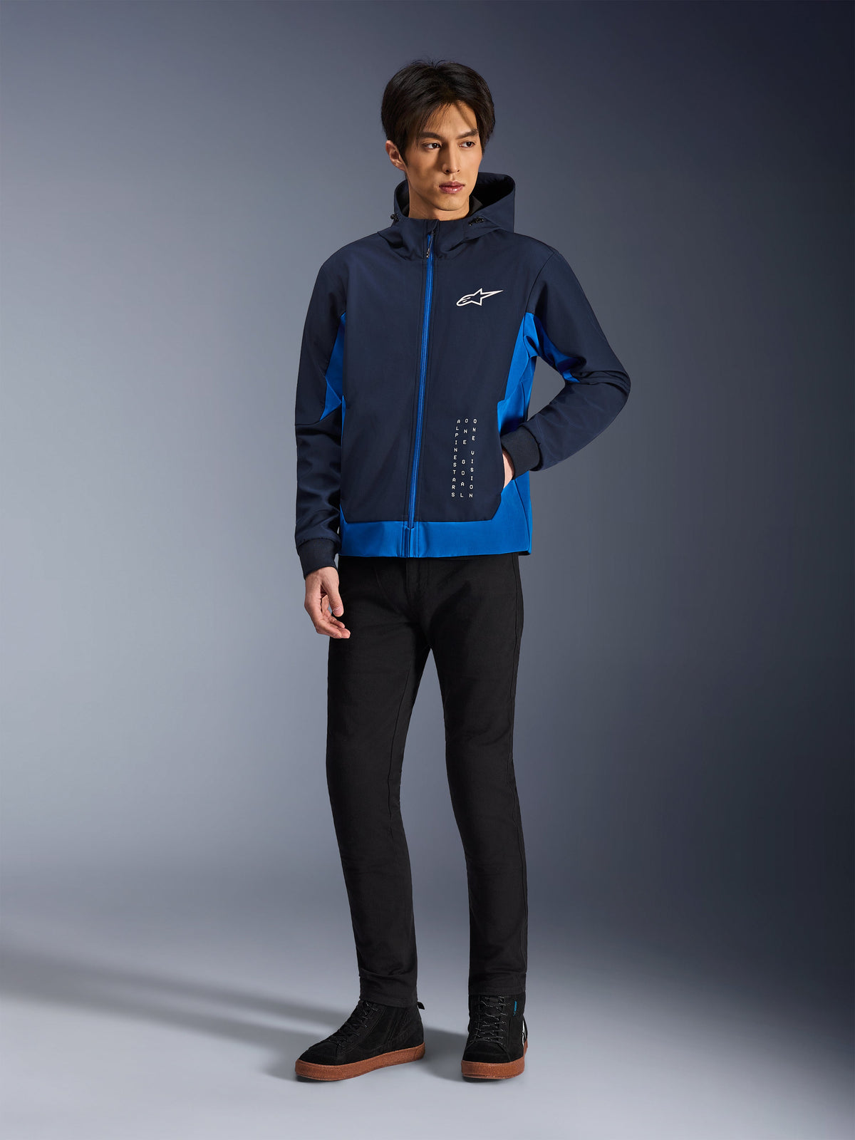 Alpinestars Radium Tech Hoodie, veste de moto urbaine, bleu marine et bleu royal, porté avec un pantalon noir et des baskets noires, avec logo Alpinestars blanc sur la poitrine et lettrage vertical, coupe athlétique décontractée pour la conduite urbaine