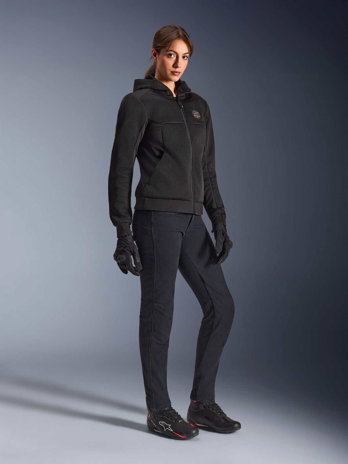 Motarde portant le hoodie Alpinestars Stella Chrome Superairflow noir, sweat à capuche technique de moto avec construction en maille 3D, associé à un pantalon de moto noir, des gants et des chaussures de moto de style sportif, esthétique sport urbain.