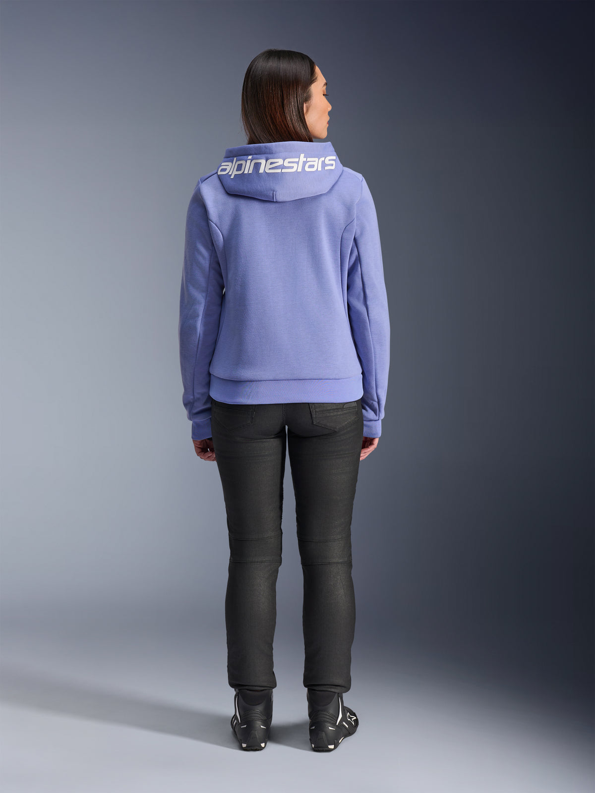Alpinestars Stella Chrome V2 Sport Hoodie, Bleu Violet, vue de dos d'une femme portant le sweat à capuche avec logo Alpinestars blanc sur la capuche, associé à un pantalon noir et des bottes de moto, vêtement de moto urbain décontracté
