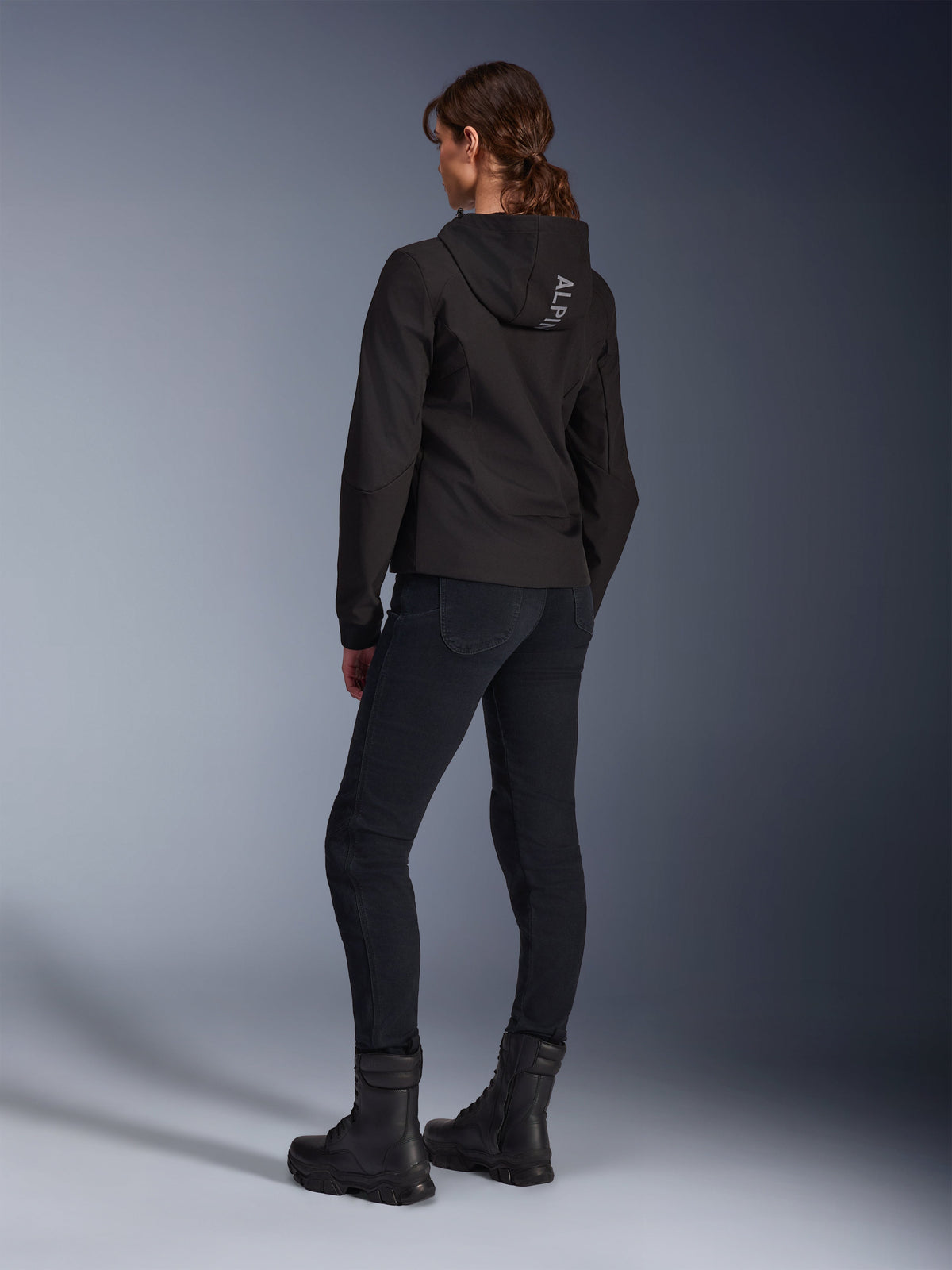 Alpinestars Stella Radium Tech Hoodie, Women's Black Urban Riding Hoodie, rear view showing 'ALPIN' branding on the hood, paired with black technical riding pants and black boots, lightweight windproof tech shell.