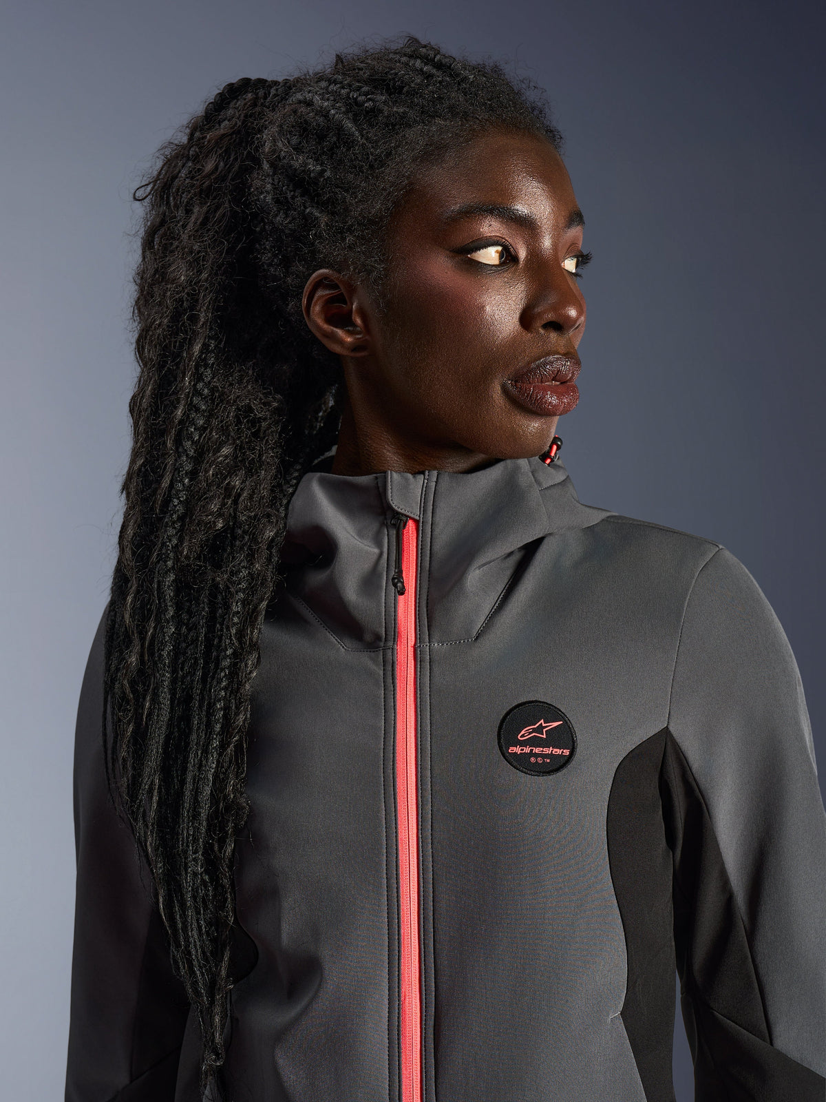 Modèle féminin portant le sweat à capuche Alpinestars Stella Radium Tech Hoodie en gris foncé et noir, avec une fermeture éclair corail vif, un logo circulaire sur la poitrine et une coque coupe-vent à col haut, tenue de moto urbaine décontractée pour femmes