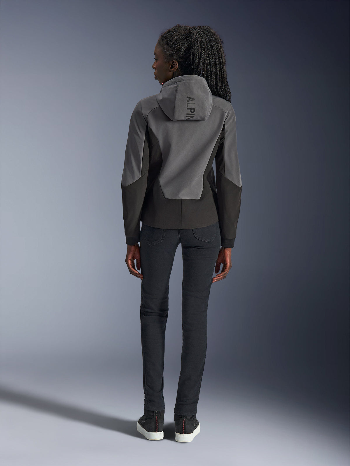 Alpinestars Stella Radium Tech Hoodie pour femme, gris foncé et noir, vue arrière montrant le marquage 'ALPIN' sur la capuche, avec un pantalon noir ajusté et des baskets noires, vêtement de moto urbain