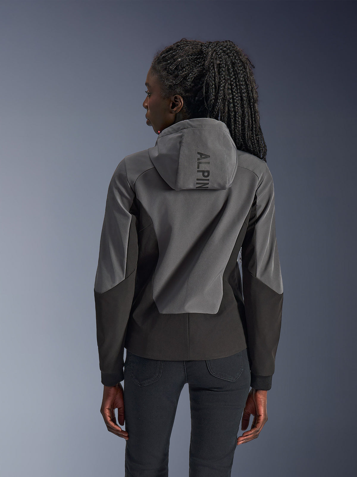 Femme portant le sweat à capuche Alpinestars Stella Radium Tech, vue de dos, Deep Gray Black, coque bicolore grise et noire à 3 couches coupe-vent, logo Alpinestars sur la capuche, vêtement de moto urbain à coupe athlétique