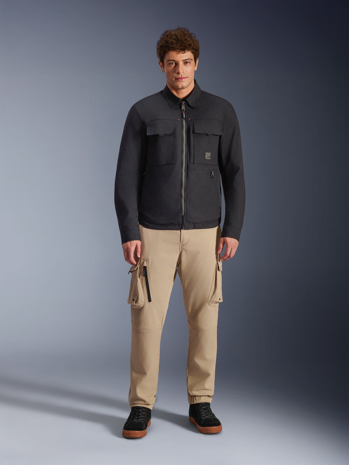 Male model wearing the Alpinestars Bryon Water Resistant Overshirt in black, featuring chest cargo pockets and a central zipper, styled with tan cargo pants and black sneakers, full-body studio portrait.