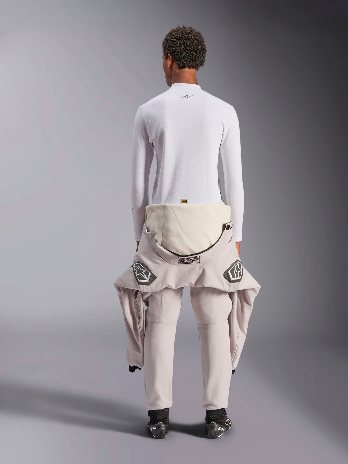 Alpinestars ZX Evo V3 Top - Long Sleeve, sous-vêtement technique de course automobile blanc, vue arrière d'une personne portant le haut ignifugé avec une combinaison rabaissée à la taille, bottines de course noires, équipement de sport automobile professionnel