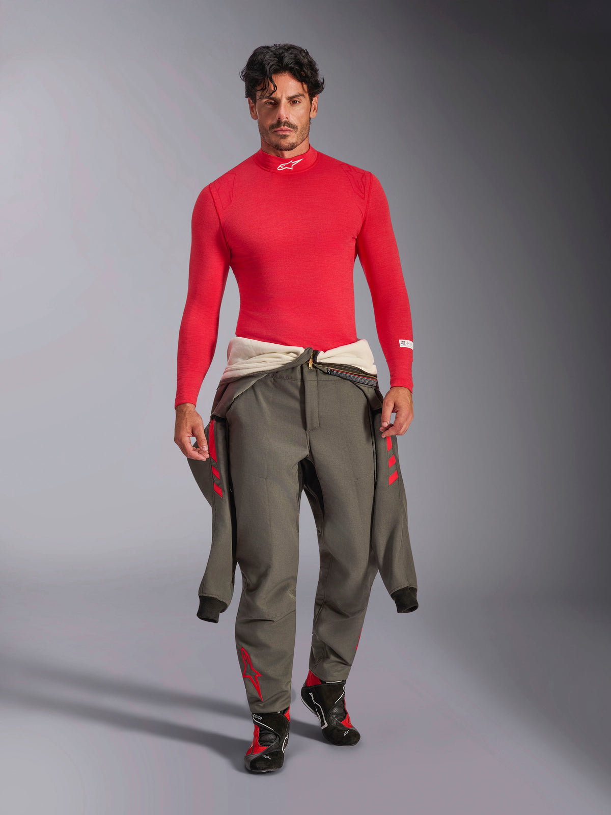 Alpinestars ZX Evo V3 Long Sleeve Top, Red Auto Base Layer, worn by a model with a grey racing suit tied at the waist and racing shoes, mock neck with white Alpinestars logo, lightweight and flame-resistant professional motorsport gear.