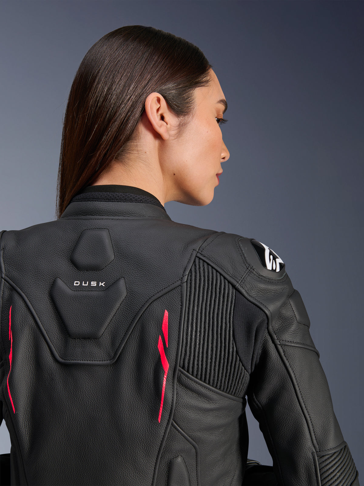 Femme portant la veste en cuir Alpinestars Stella Dusk, cuir noir avec des accents rose Diva et logo blanc sur l'épaule, vue arrière montrant le marquage DUSK et les panneaux extensibles en accordéon, design ergonomique sportif