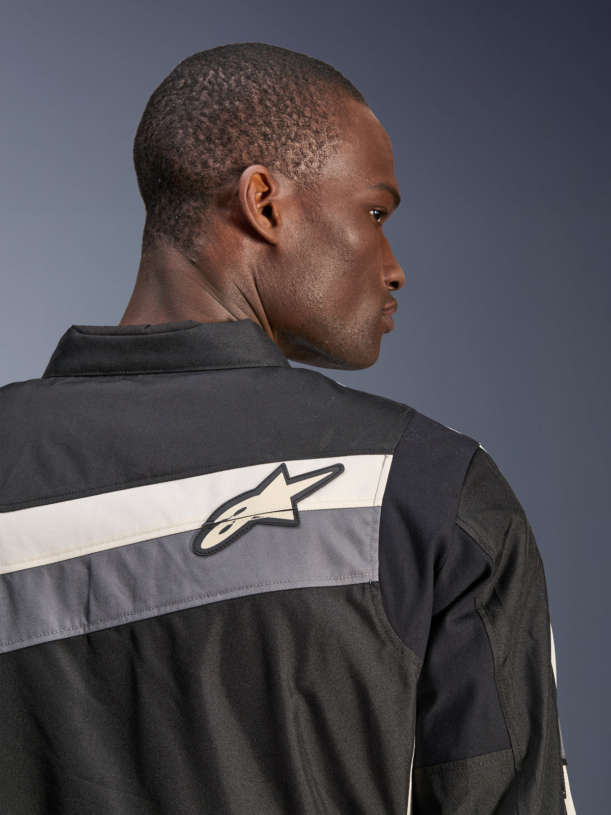 Veste Alpinestars T-Dyno WR, veste de moto en textile, noir, gris foncé et écru, gros plan du haut du dos et de l'épaule avec bandes horizontales et logo étoile Alpinestars, portée par un mannequin, design d'inspiration vintage pour la route