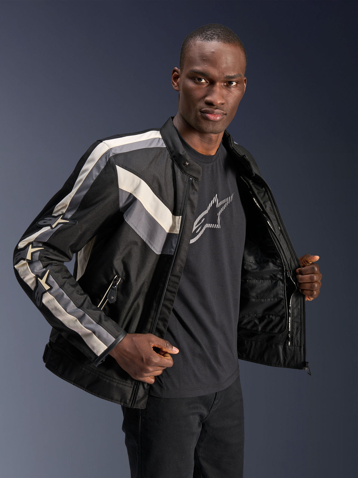 Veste Alpinestars T-Dyno WR, noir, gris foncé et écru, veste de moto en textile avec coupe biker d'inspiration vintage, détails à rayures sur la poitrine et les manches, portée sur un t-shirt graphique noir, équipement de moto urbain
