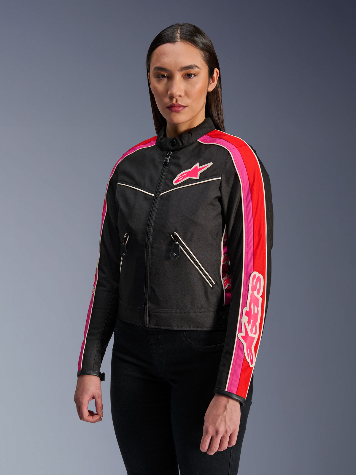 Femme portant la veste Alpinestars Stella T-Dyno WR en Noir Rouge Éclatant Fuchsia, veste de moto en textile avec une coupe biker d'inspiration vintage, présentant des rayures fuchsia et rouges éclatantes sur les manches et un logo sur la poitrine, associée à un pantalon noir.