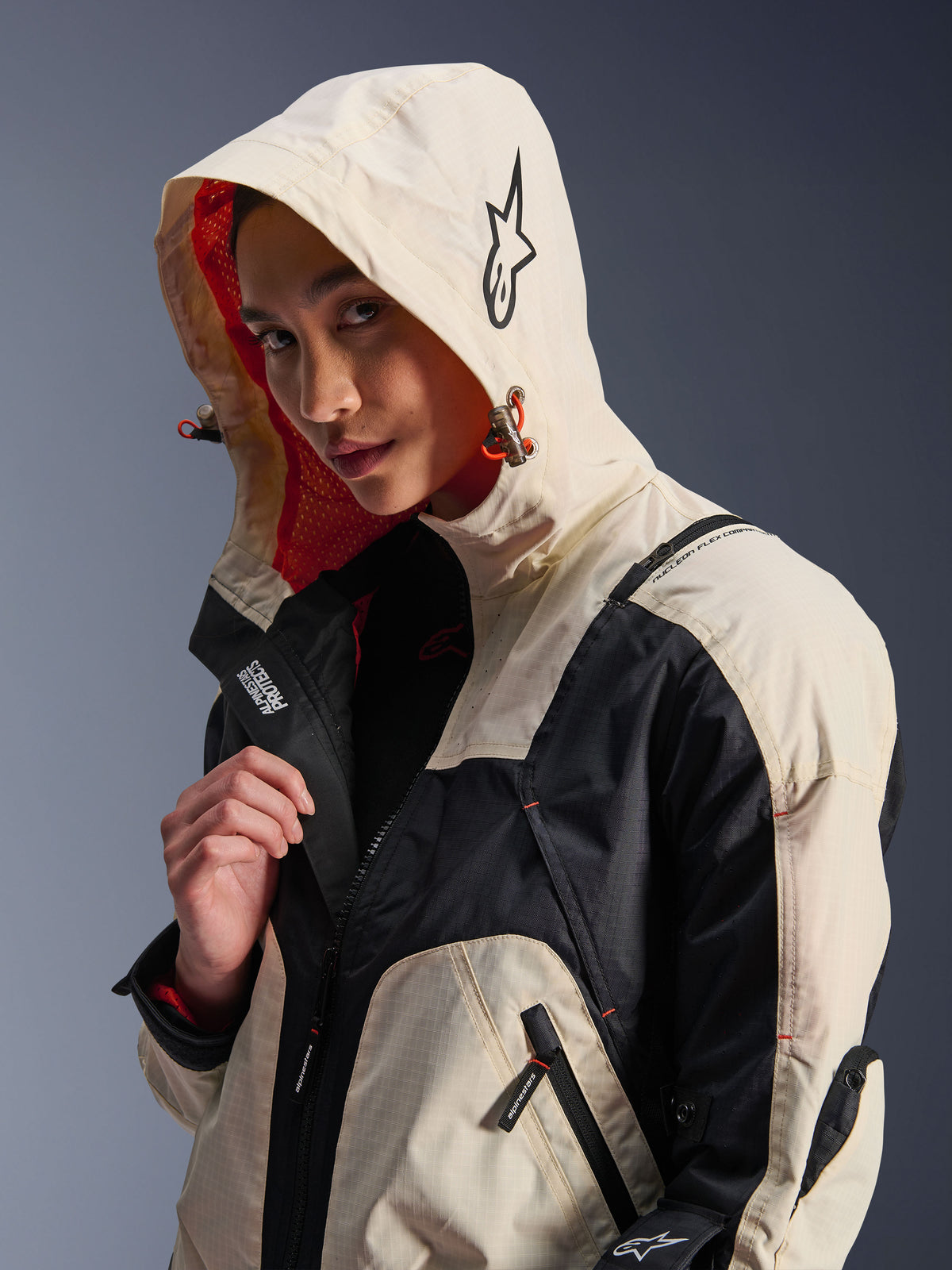 Veste Alpinestars Molly Air en Black Almond, portée par un mannequin femme avec la capuche relevée, présentant un design color-block crème et noir, une doublure en mesh orange vif et des éléments de design urbain technique incluant un logo étoile et des détails de zips.