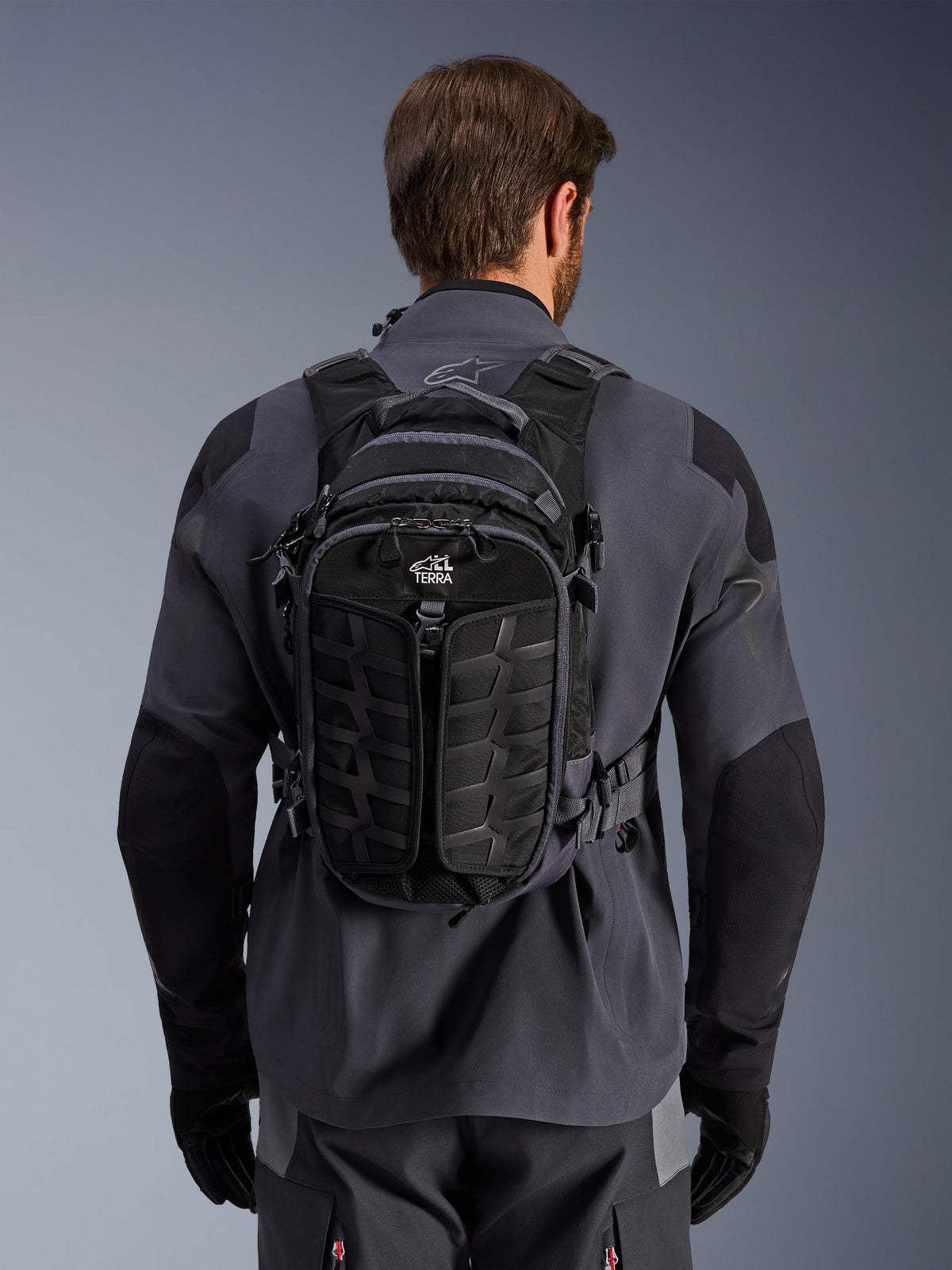 Sac à dos Alpinestars AT-15, sac à dos de moto d'aventure, noir et gris foncé, porté par une personne sur une veste de moto technique grise, vue arrière montrant le marquage « ALL TERRA », design ergonomique avec panneaux texturés renforcés et sangles de maintien.
