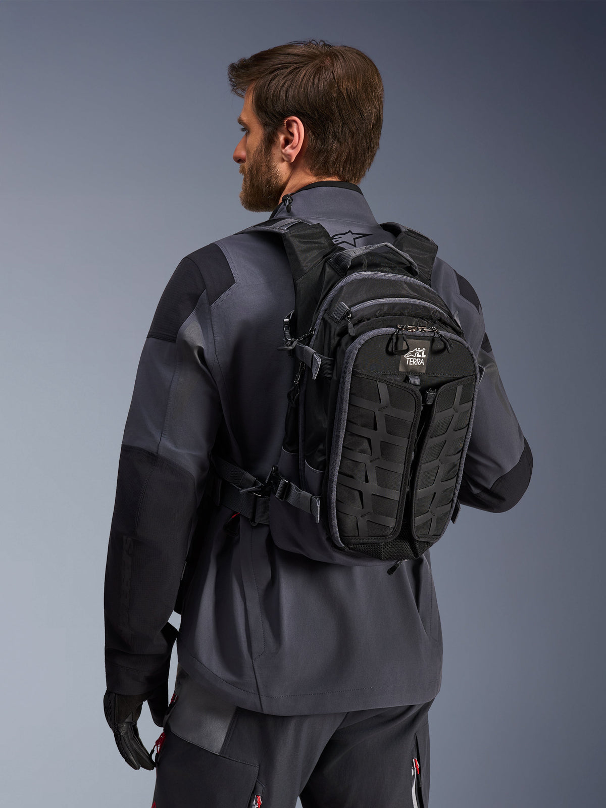 Sac à dos Alpinestars AT-15, sac à dos de moto aventure, noir et gris foncé, porté par un pilote en veste grise, doté d'un design technique avec des sangles MOLLE et le marquage « TERRA », équipement tout-terrain ergonomique
