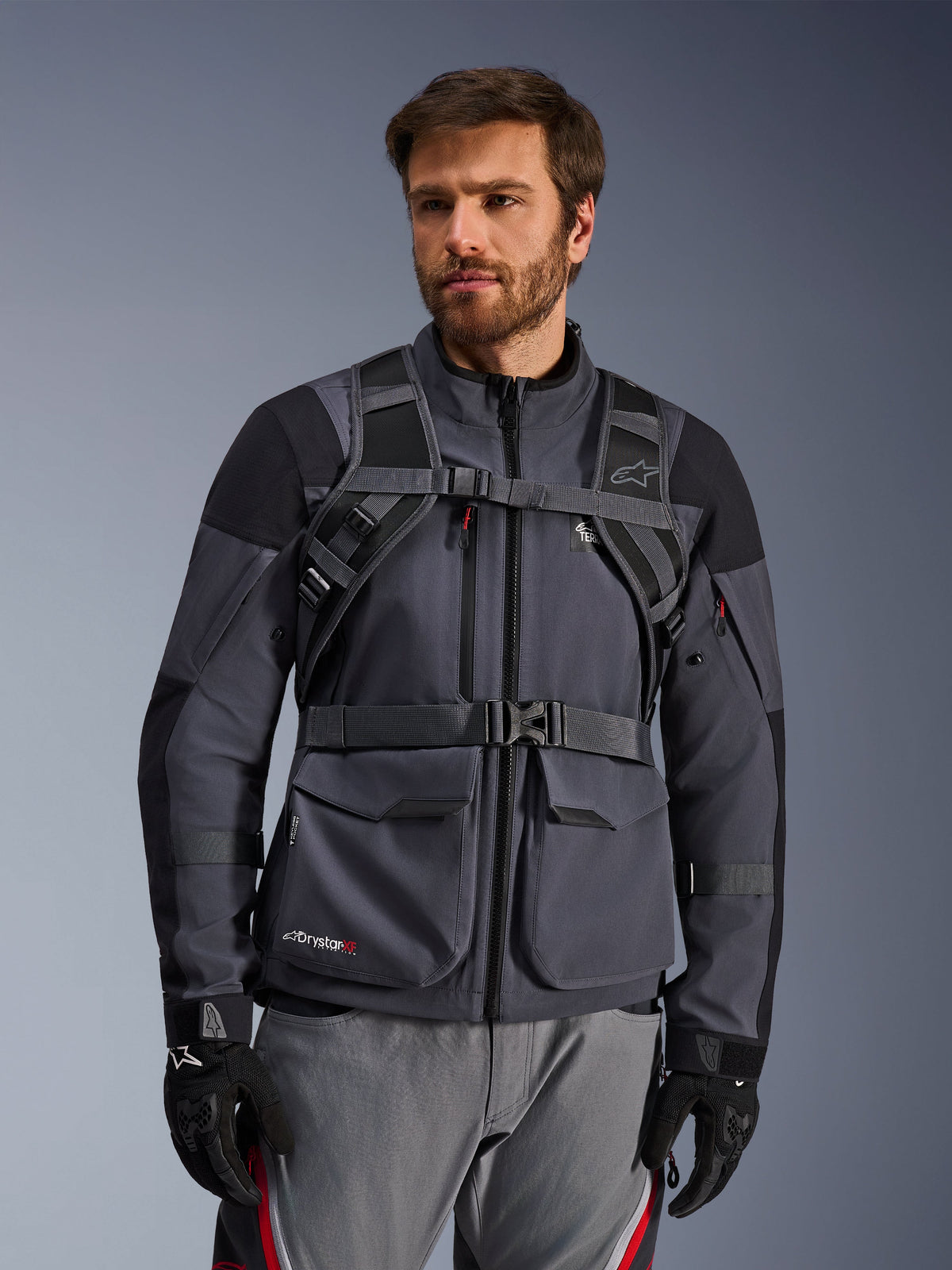 Système de harnais du sac à dos Alpinestars AT-15, Noir Deep Gray, vue de face porté par un pilote sur une veste de moto grise, avec fermeture magnétique de poitrine et sangle ventrale sécurisée, associé à des gants noirs pour la moto d'aventure