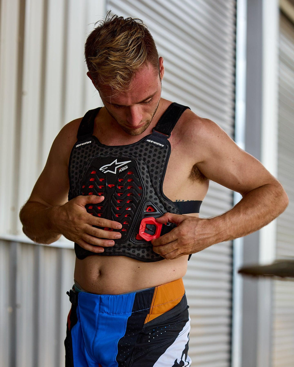 Homme enfilant le pare-pierres Alpinestars A-5 Plasma, noir avec accents rouges et logo blanc, structure en maille nid d'abeille, porté avec un pantalon de motocross bleu et orange, préparatifs pour une course tout-terrain