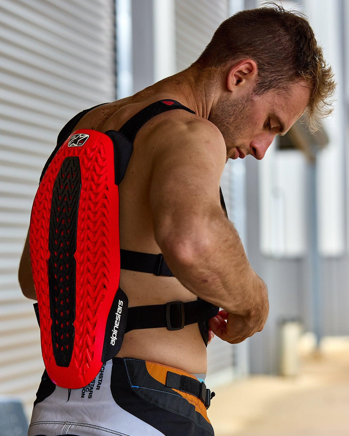 Pare-pierres Alpinestars A-5 Plasma, rouge et noir, doté d'une protection dorsale texturée avec technologie Nucleon PLASMA, porté par un homme ajustant des sangles noires, vue de profil mettant en avant la coque protectrice et la conception flexible pour la conduite tout-terrain