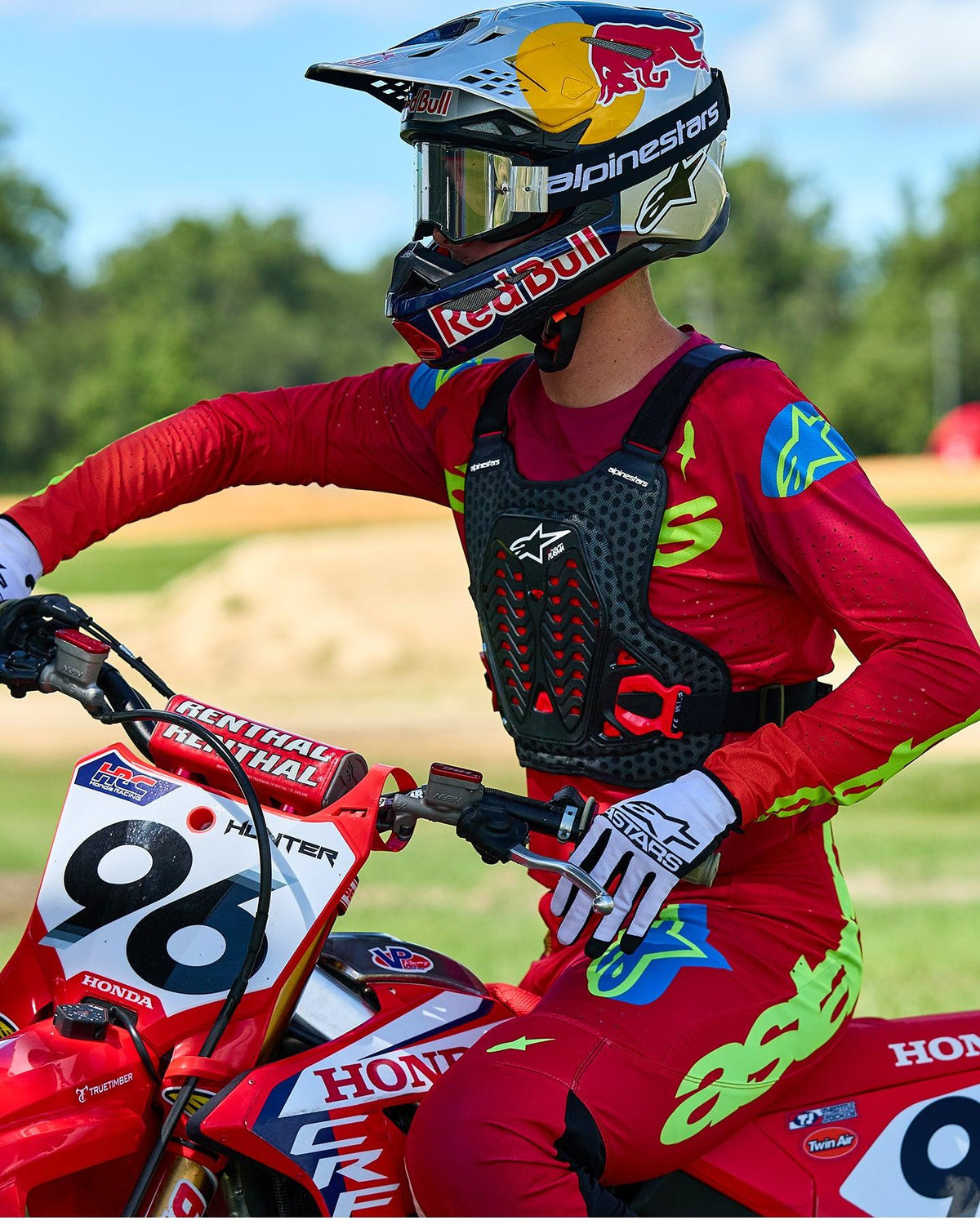 Pilote de motocross portant le pare-pierres Alpinestars A-5 Plasma noir et rouge, avec un maillot et un pantalon de course rouge et vert citron, assis sur une moto Honda rouge, équipement de protection tout-terrain haute performance