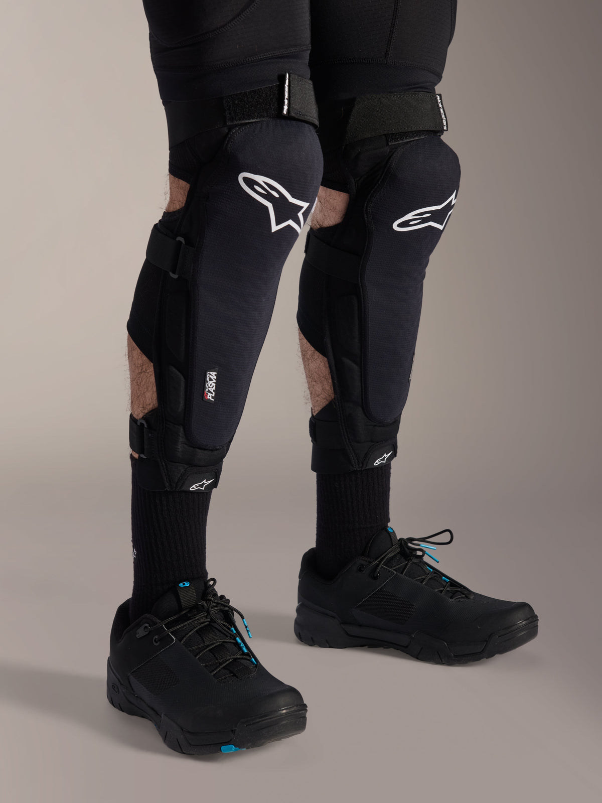 Genouillères Alpinestars A-Impact Plasma Pro, noir et blanc, avec le marquage de la technologie Nucleon PLASMA, portées avec des chaussettes noires et des chaussures de VTT noires, sangles à crochet et boucle sécurisées, équipement de protection ergonomique pour le cyclisme tout-terrain