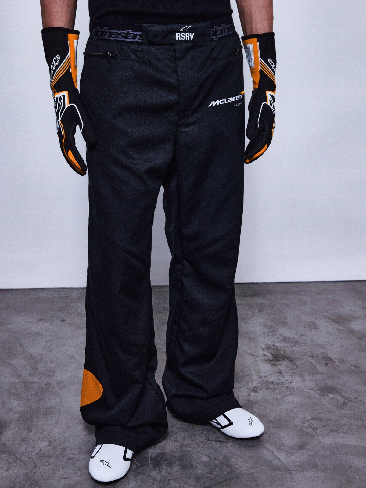 Alpinestars RSRV Arena Suit Pants, pantalon de course noir avec logos McLaren et Alpinestars, détails orange, porté avec gants noirs et orange et chaussures de course blanches