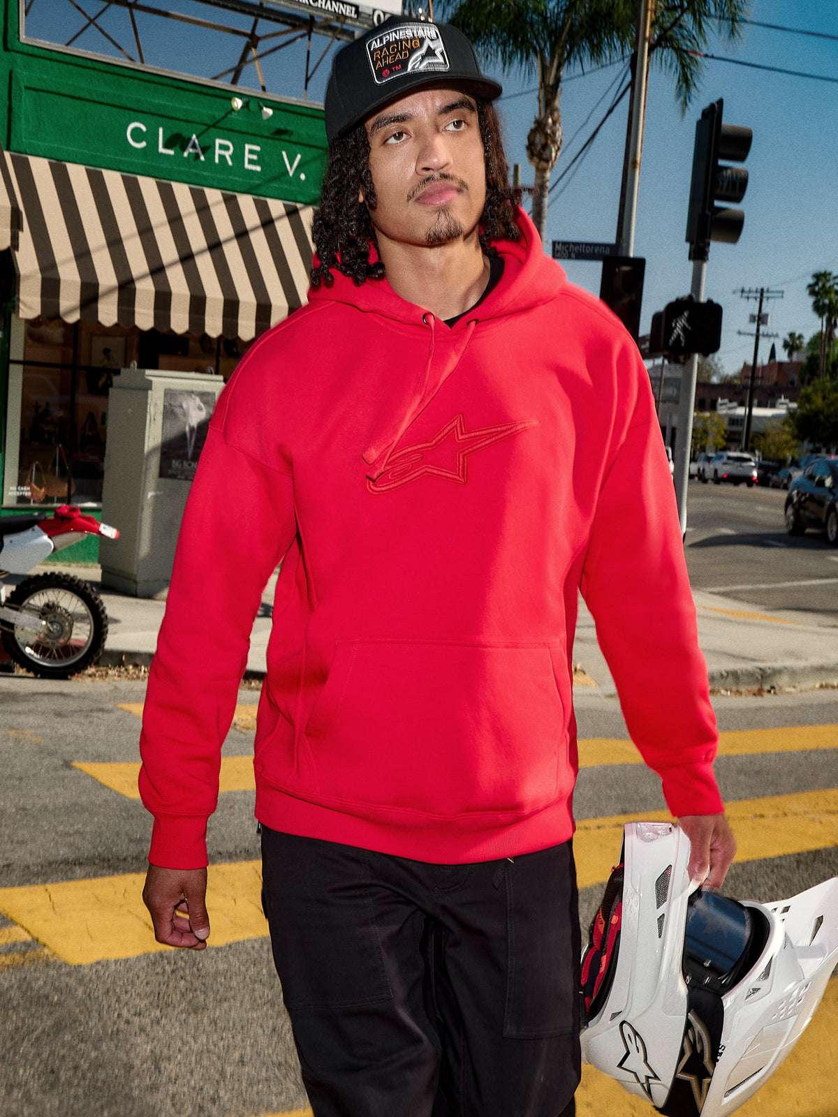 Sweat à capuche Emergent Oversized Alpinestars, rouge, logo Astars brodé en 3D sur la poitrine, porté avec un pantalon noir et une casquette de baseball tout en tenant un casque de motocross blanc, style streetwear urbain décontracté