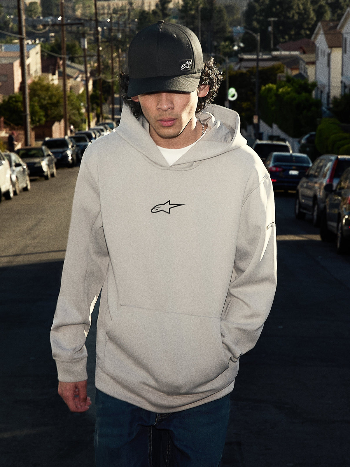 Sweat à capuche Alpinestars Insight, streetwear décontracté, gris ciment avec logo Astars noir sur la poitrine, style à enfiler avec poche kangourou, porté avec une casquette noire dans une rue résidentielle, design moderne inspiré du sport automobile