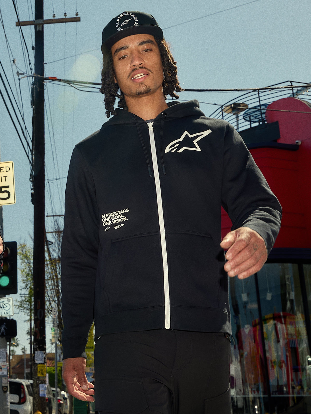 Sweat à capuche Transverse Alpinestars, polaire noire à fermeture éclair frontale, fermeture éclair blanche et logo Astars blanc sur la poitrine, impression du texte « One Goal One Vision », style streetwear décontracté, porté par un homme dans un environnement urbain extérieur