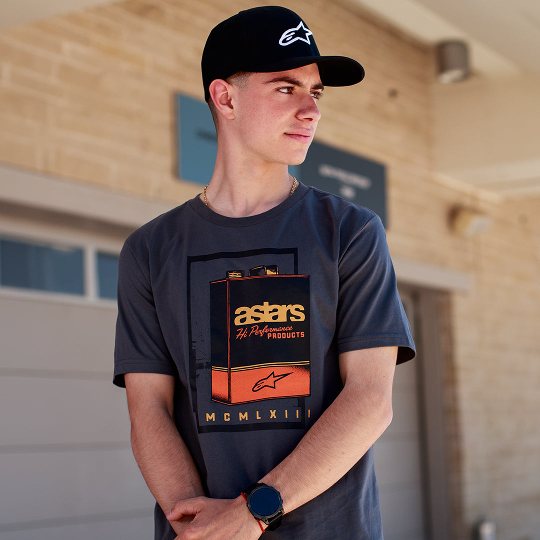 T-shirt Alpinestars Galun CSF, t-shirt à manches courtes, gris, motif graphique de bidon d'essence vintage à l'avant avec marquage « astars » et chiffres romains « MCMLXIII », porté par un homme avec une casquette noire à logo, style motorsport décontracté