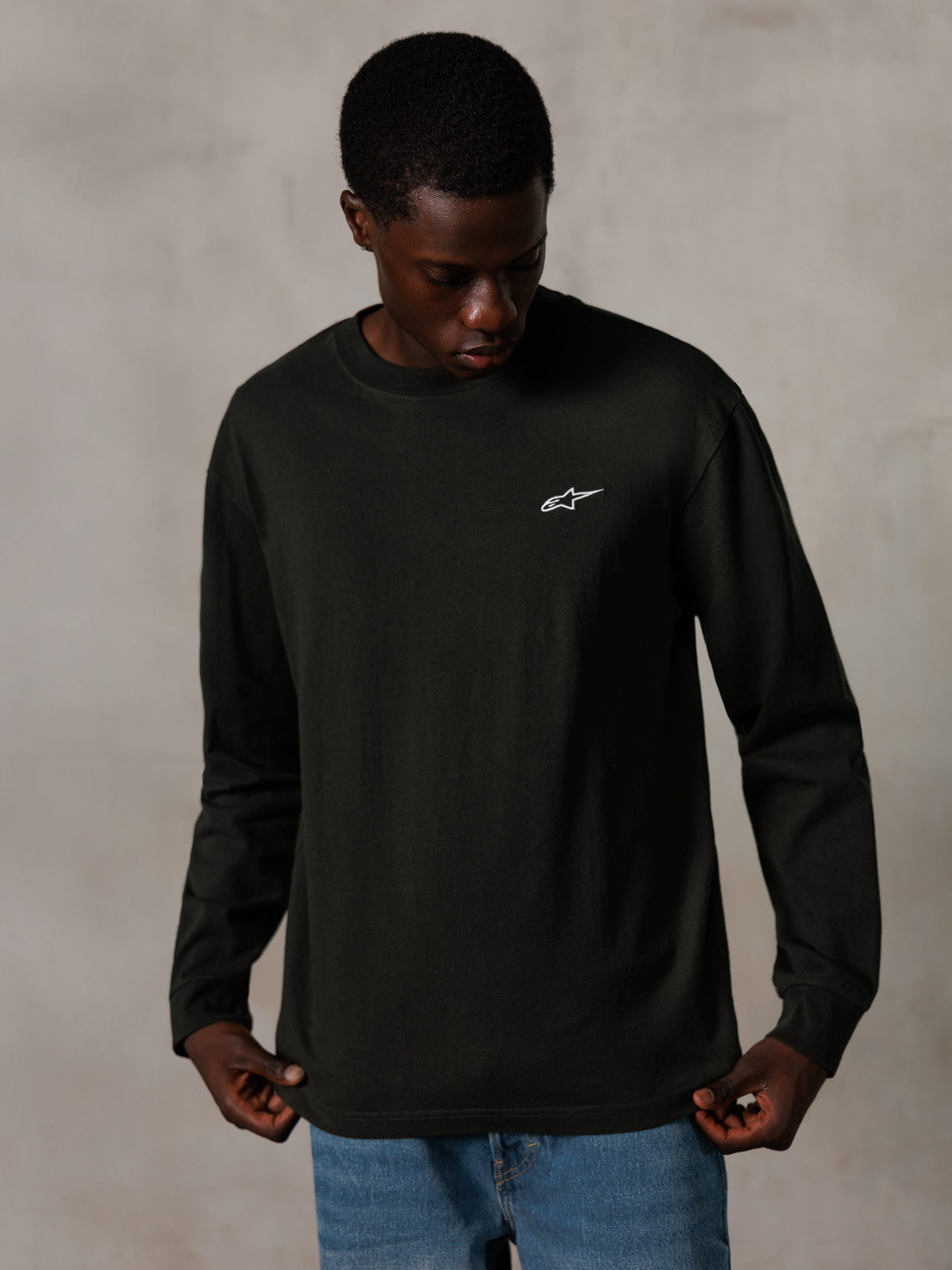Alpinestars Essentials Ageless LS Tee, t-shirt à manches longues, noir délavé, arborant un petit logo blanc sur la poitrine, porté par un mannequin avec un jean bleu, style streetwear décontracté