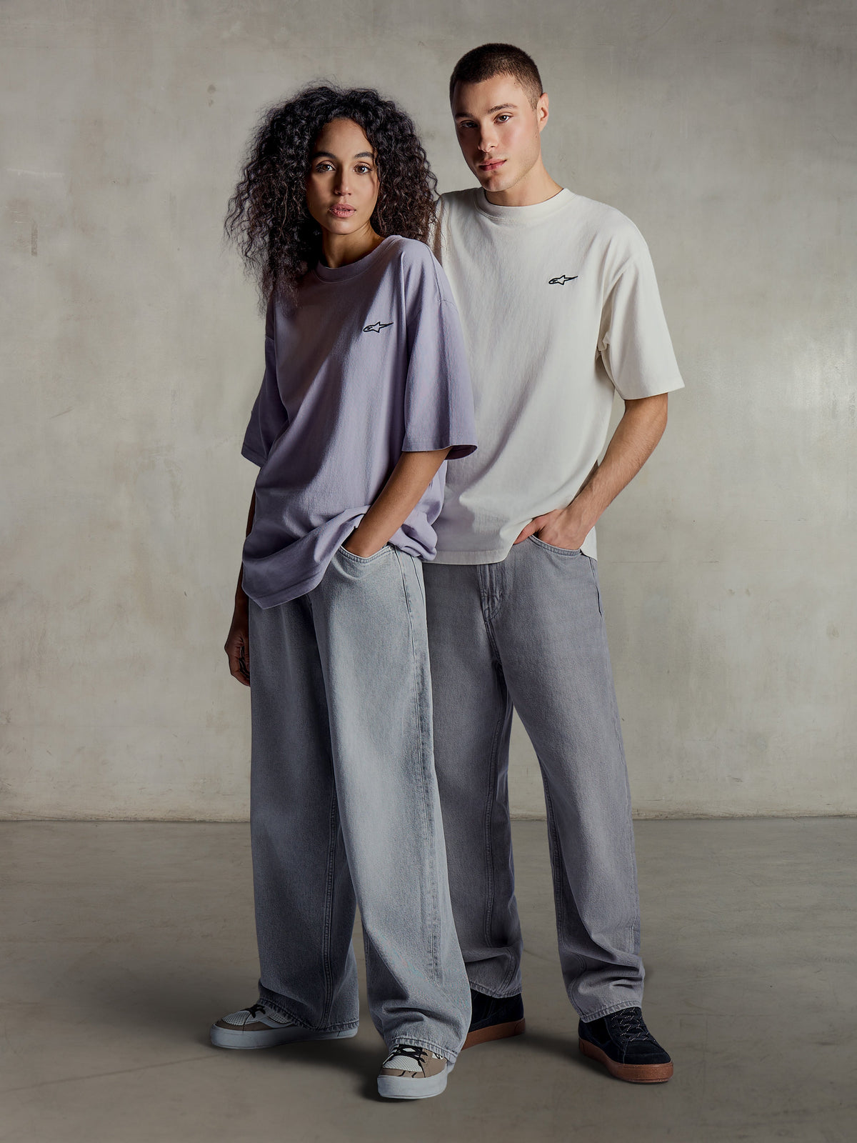 Alpinestars Essentials Ageless SS Tee, vue lifestyle de deux modèles portant des t-shirts à manches courtes en violet clair et argenté, coton biologique à coupe décontractée avec un logo discret sur la poitrine, associés à des jeans larges pour un style streetwear décontracté.