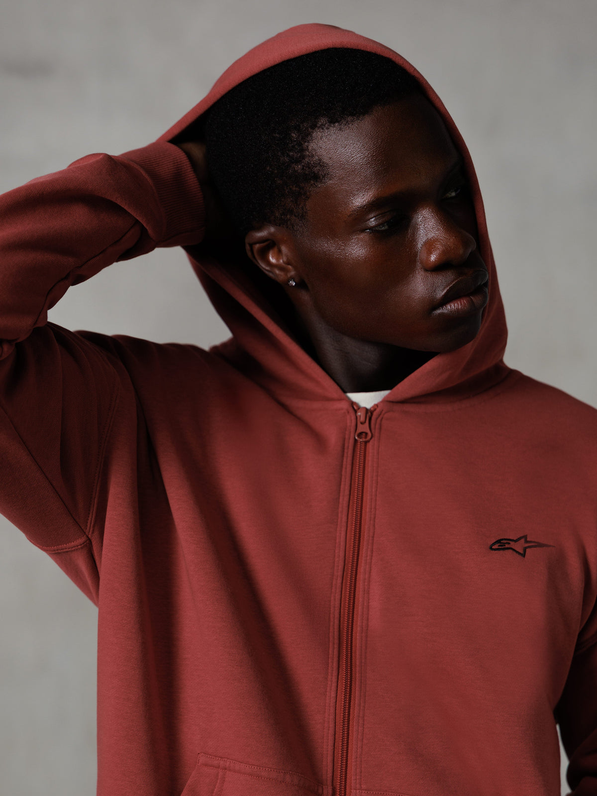 Sweat à capuche zippé Essentials Ageless Alpinestars, rouge brique, streetwear décontracté, capuche relevée avec la main derrière la tête, petit logo Astars noir sur la poitrine, sweat en polaire à coupe décontractée