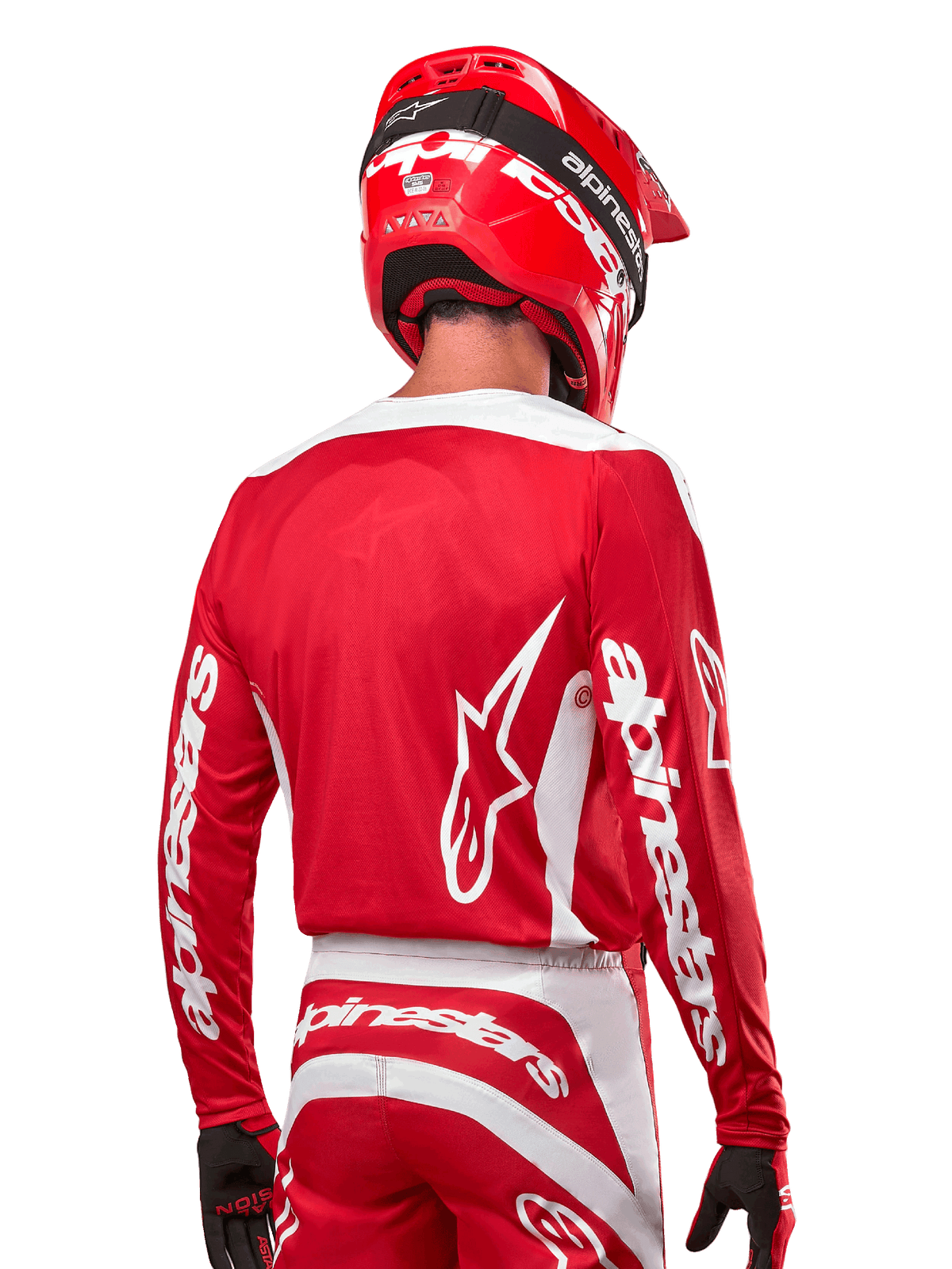 Pilote de motocross portant le maillot Alpinestars Fluid Lurv 2024, rouge Mars et blanc, vue arrière montrant le logo Alpinestars blanc au dos et le marquage sur les manches, associé à un casque rouge et des gants noirs, équipement de course tout-terrain professionnel