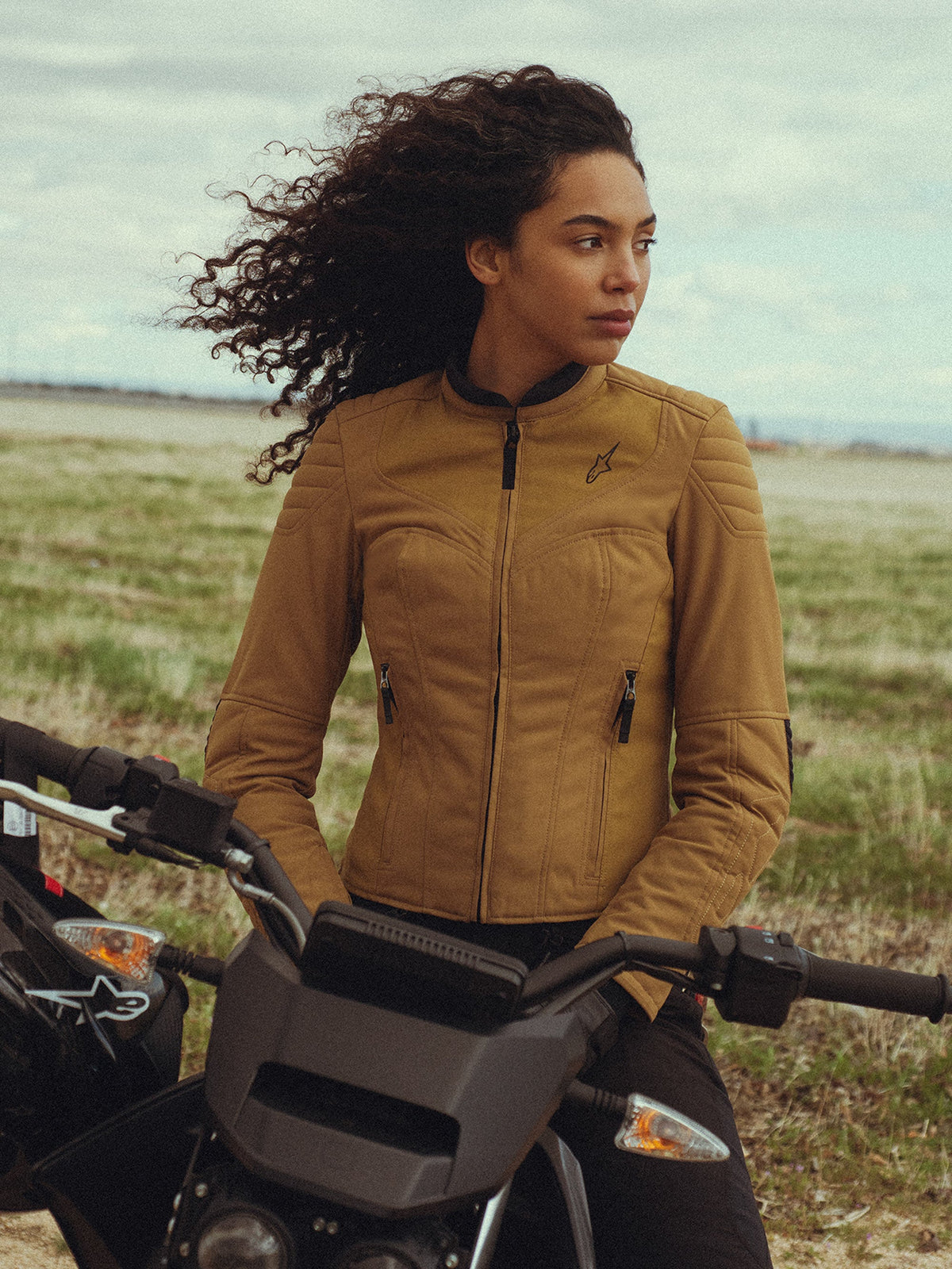 Veste Alpinestars Isla WR pour femme en Utility Brown Light Gold, portée par une femme assise sur une moto dans un champ en extérieur, avec des détails matelassés aux épaules, une silhouette ajustée et un style urbain contemporain.