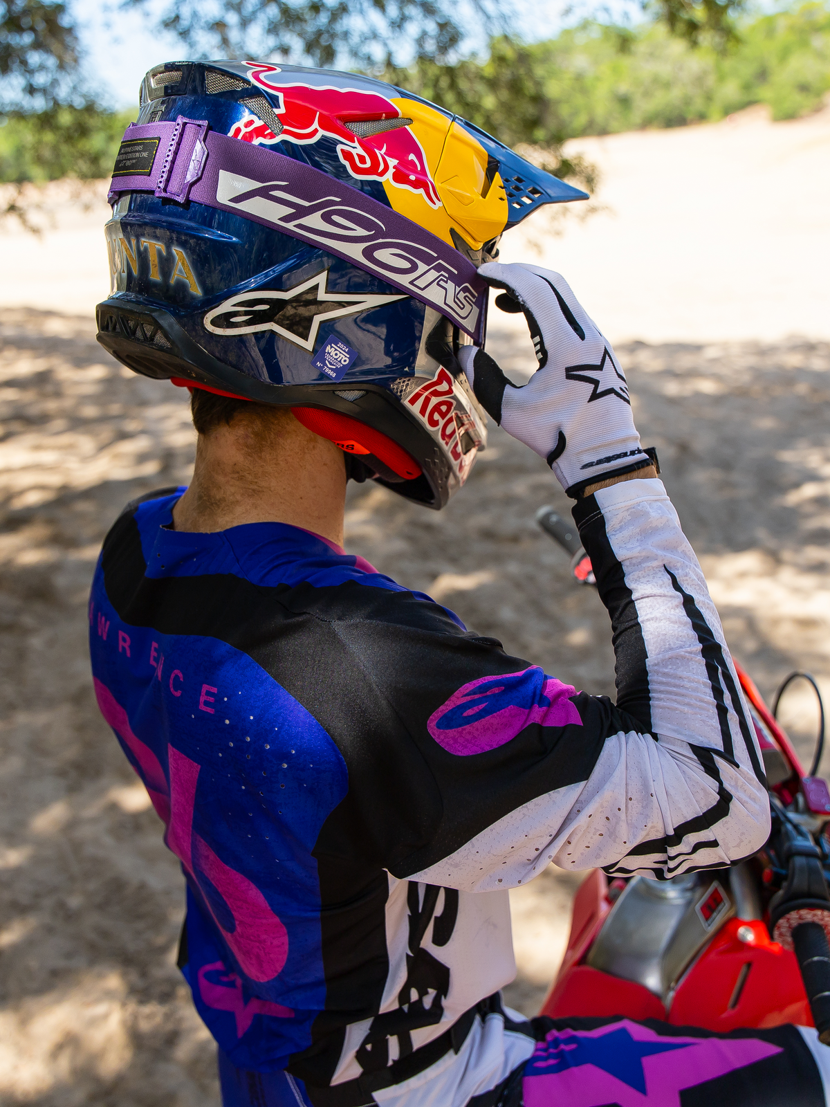 Le pilote de motocross Hunter Lawrence portant le masque Supertech Vision HUNTA96 XXV en édition limitée, bandeau violet avec 'H96' et coutures jaunes, casque Red Bull bleu, maillot violet et blanc avec accents noirs, gants blancs, ajustant son masque dans un décor tout-terrain sablonneux