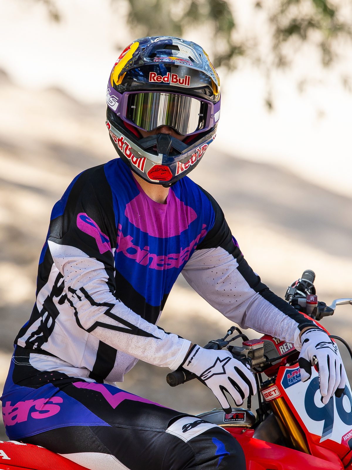 Pilote de motocross sur une moto tout-terrain rouge, masque Supertech Vision HUNTA96 XXV Alpinestars en édition limitée violet et blanc avec verres miroir argentés, maillot et pantalon Alpinestars assortis violet, noir et blanc, casque Red Bull, gants blancs, équipement de course professionnel coordonné.