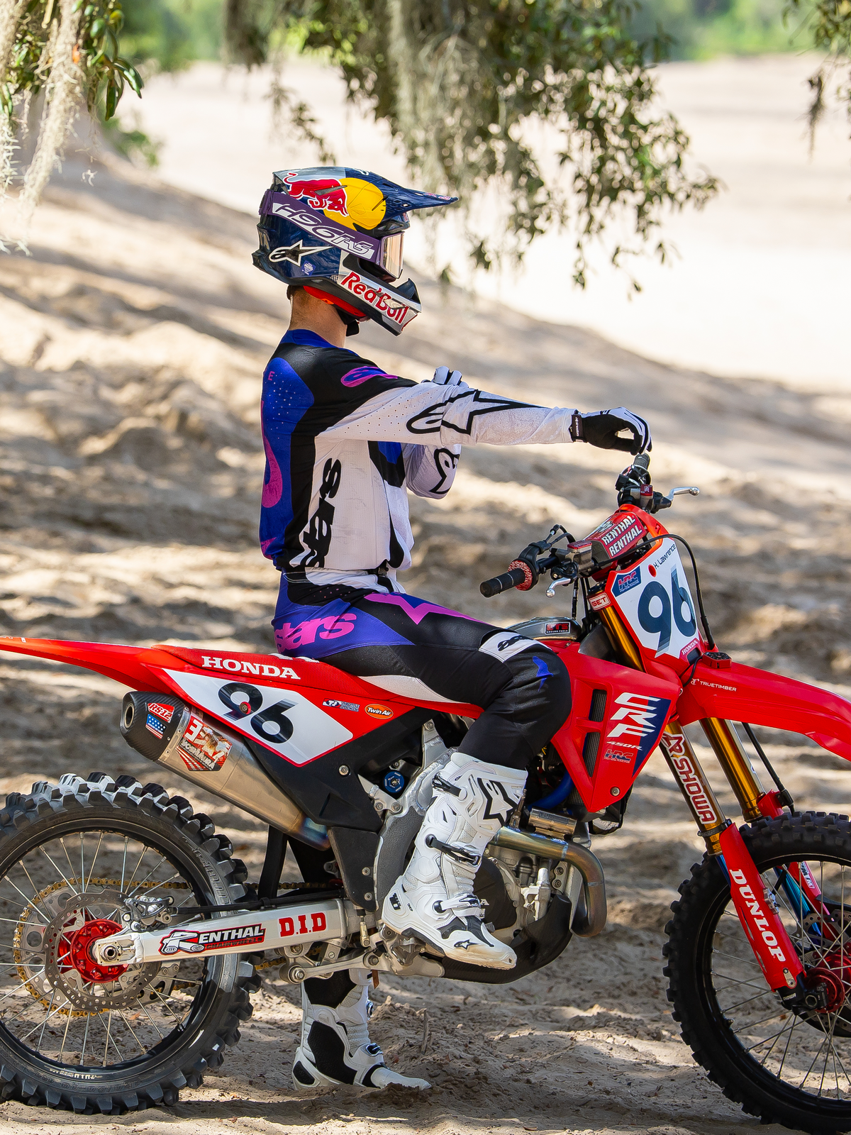 Pilote de motocross portant le masque Limited Edition HUNTA96 XXV Supertech Vision en Violet Blanc Miroir Argent, maillot de course assorti violet et blanc et bottes blanches, assis sur une moto tout-terrain Honda rouge avec le numéro 96, dans un cadre tout-terrain extérieur sur une piste sablonneuse.