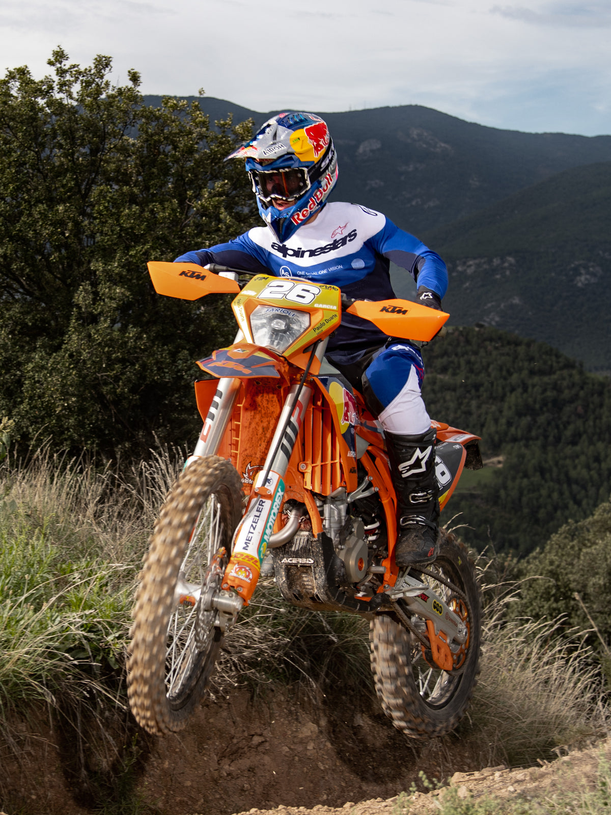 Motocross rider in action on an orange KTM dirt bike, wearing Alpinestars Pro-Dura gear in Navy, Bright Blue, and White. Professional off-road outfit including a blue and white jersey and matching pants, worn with a Red Bull helmet and black boots on a mountain trail.
