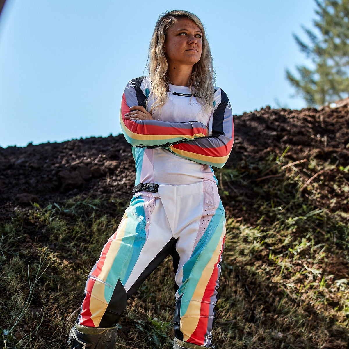 Pilote de motocross femme portant le pantalon Alpinestars Stella Techstar 2024 et le maillot assorti, design Noir Multicolore avec base blanche et rayures éclatantes bleues, jaunes et rouges, debout les bras croisés sur une piste de terre, tenue de course tout-terrain professionnelle