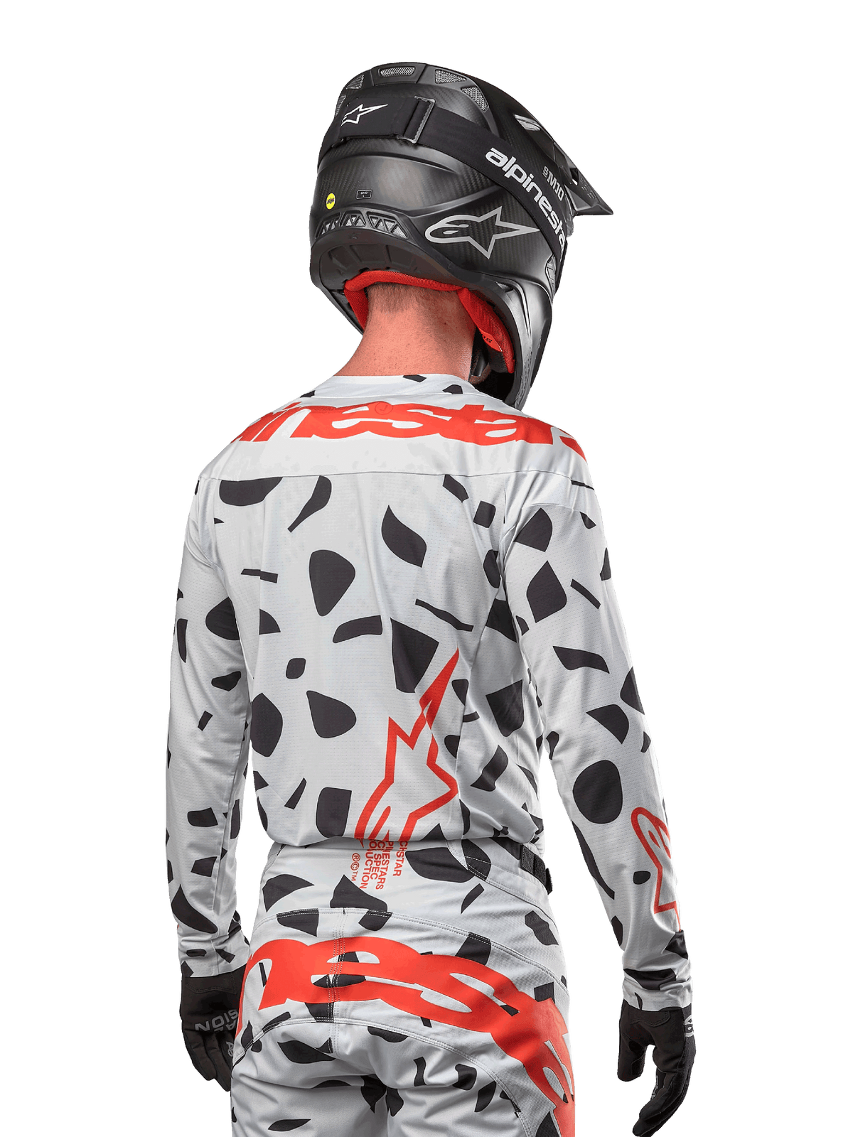 Pilote de motocross vu de dos portant le maillot Alpinestars 2024 Techstar Rantera en Haze Gray Camo, doté d'une base gris clair avec des taches graphiques noires et des logos Alpinestars rouges éclatants sur le dos et les épaules, associé à un pantalon assorti et un casque noir.