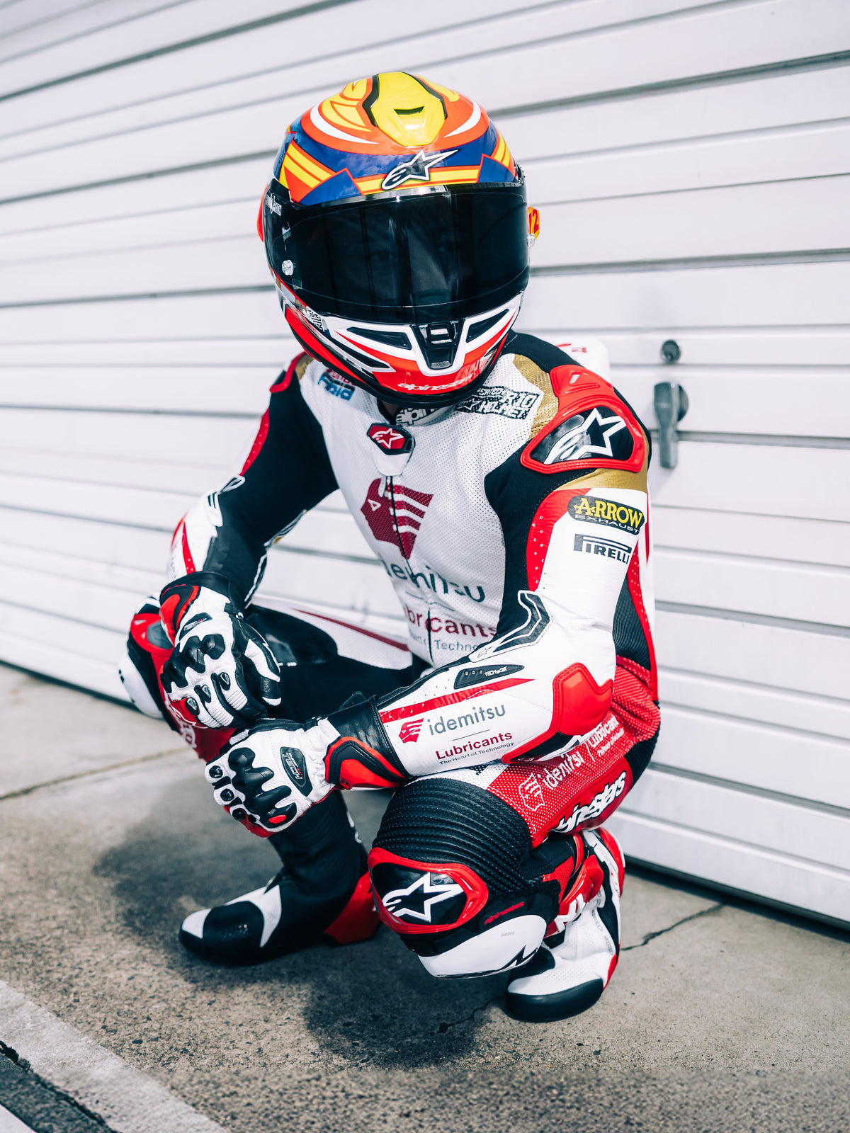 Casque Alpinestars S-R7 Taiyo Furusato, casque moto intégral racing, rouge, blanc, bleu et jaune fluo, certifié ECE 22.06, porté par un pilote de moto en combinaison en cuir de course avec écran teinté.