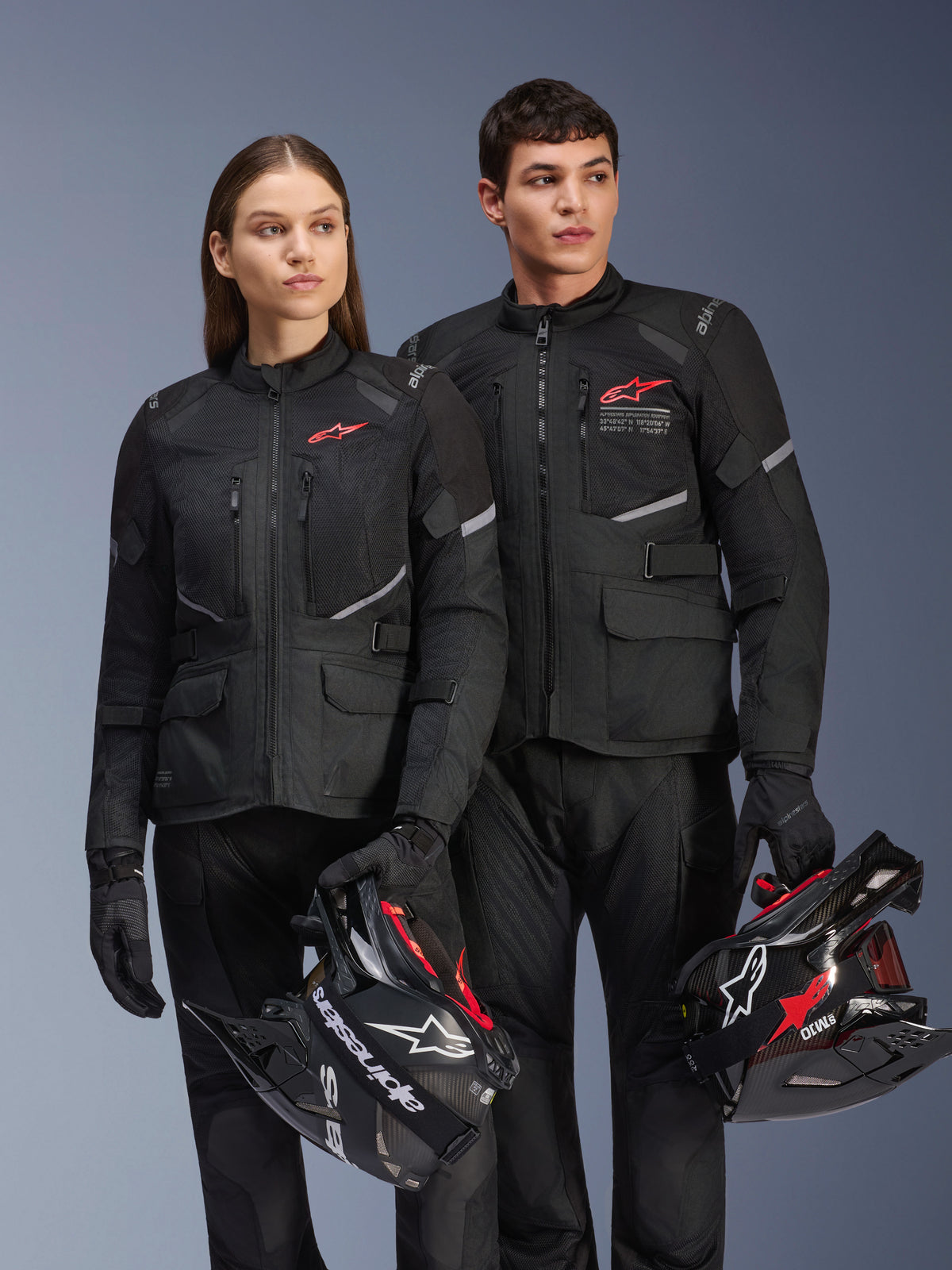 Homme et femme portant des vestes Alpinestars Andes Air Drystar®, équipement de touring aventure noir avec panneaux en mesh et détails réfléchissants, pantalons et gants noirs assortis, tenant des casques de motocross, photo de studio de vêtements de moto professionnels