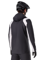 Veste de pluie Alpinestars A-Dura, veste de pluie pour VTT, noire avec empiècements blancs sur les manches, vue arrière d'un cycliste portant un casque de VTT noir et des gants, construction technique imperméable à 2,5 couches, équipement de VTT