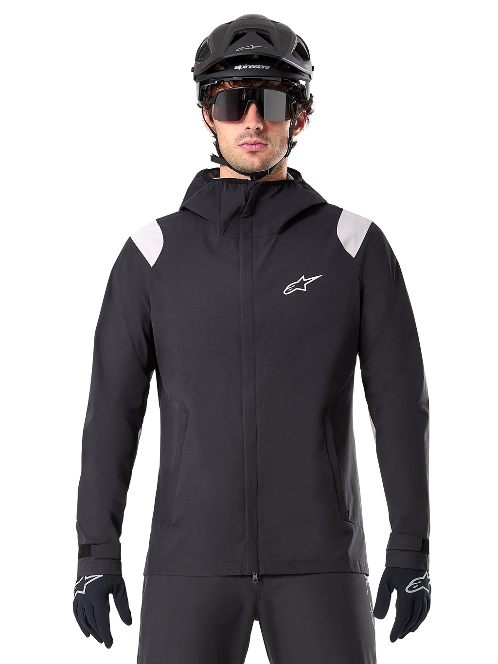 Veste de pluie Alpinestars A-Dura, veste de pluie VTT, noire avec empiècements blancs aux épaules, portée avec un casque de VTT noir, des lunettes de soleil et des gants, conception imperméable avec capuche et fermeture éclair frontale, équipement de sport de plein air pour le cyclisme