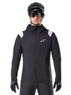 Veste de pluie Alpinestars A-Dura, veste de pluie VTT, noire avec empiècements blancs aux épaules, portée avec un casque de VTT noir, des lunettes de soleil et des gants, conception imperméable avec capuche et fermeture éclair frontale, équipement de sport de plein air pour le cyclisme