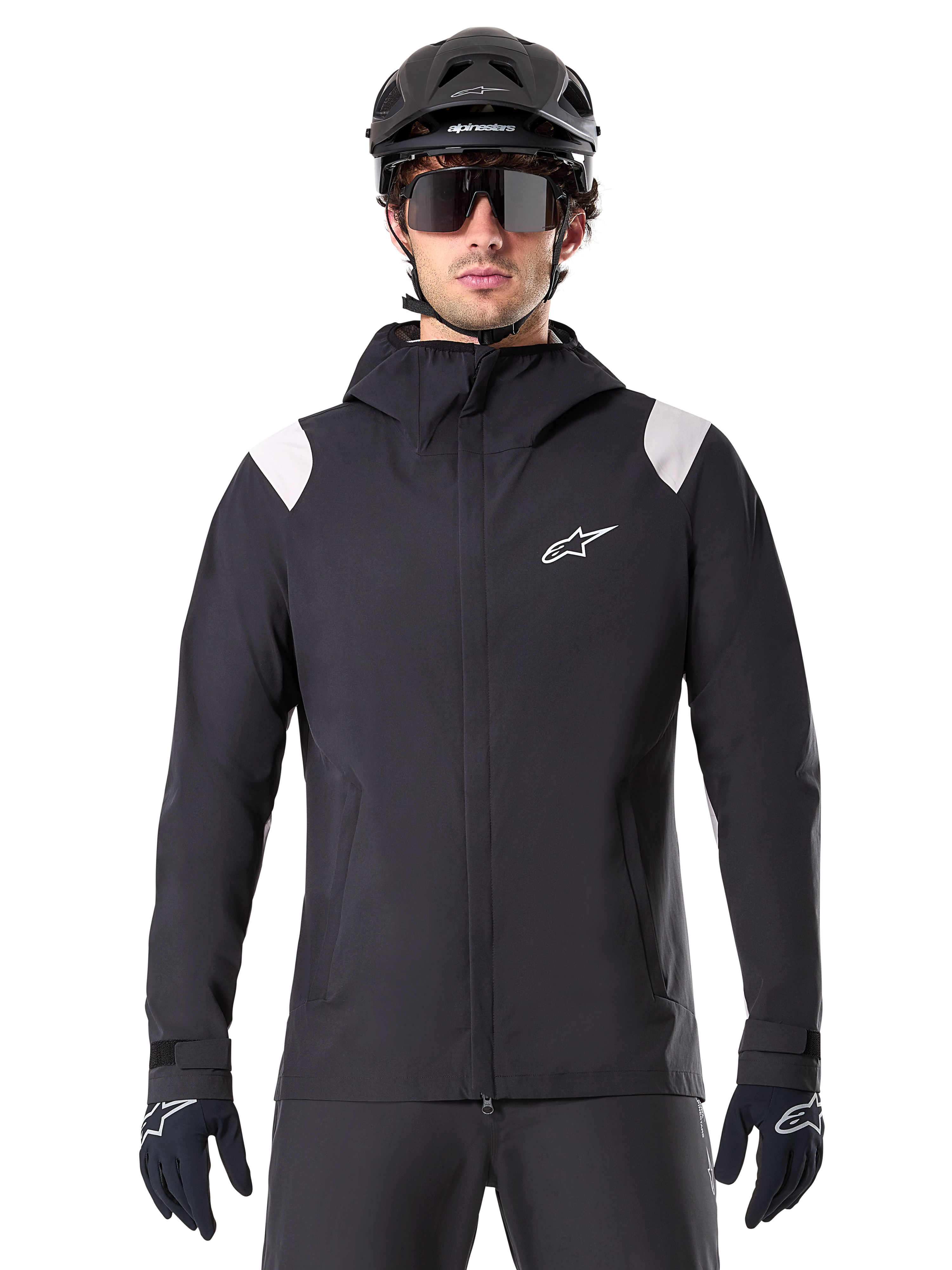 Veste de pluie Alpinestars A-Dura, veste de pluie VTT, noire avec empiècements blancs aux épaules, portée avec un casque de VTT noir, des lunettes de soleil et des gants, conception imperméable avec capuche et fermeture éclair frontale, équipement de sport de plein air pour le cyclisme